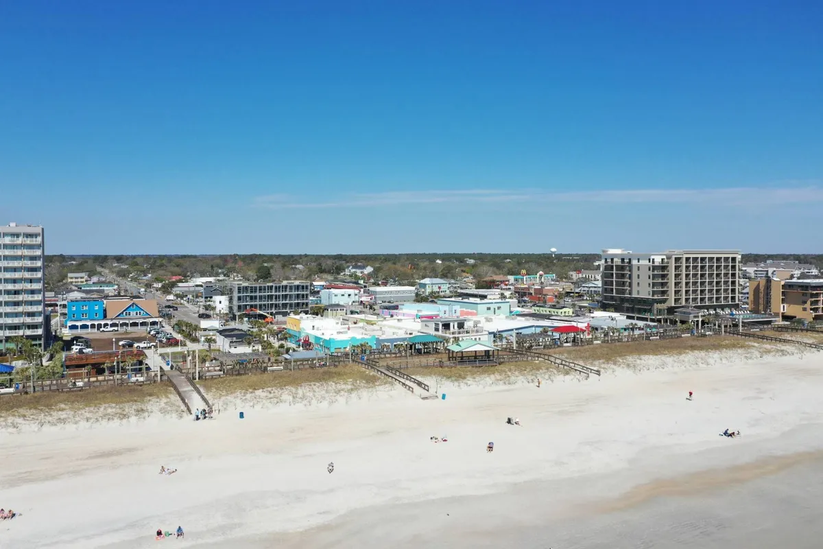 A lively exterior shot featuring the Carolina Beach boardwalk with colorful buildings, beachfront views, and people enjoying the vibrant local scene. The atmosphere is energetic, hinting at nearby restaurants, bars, and local festivities.