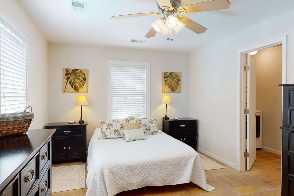 A cozy bedroom showcasing a bed with decorative pillows, nightstands with lamps on either side, and palm leaf wall art. The soft lighting and neutral tones evoke a sense of peace and relaxation.