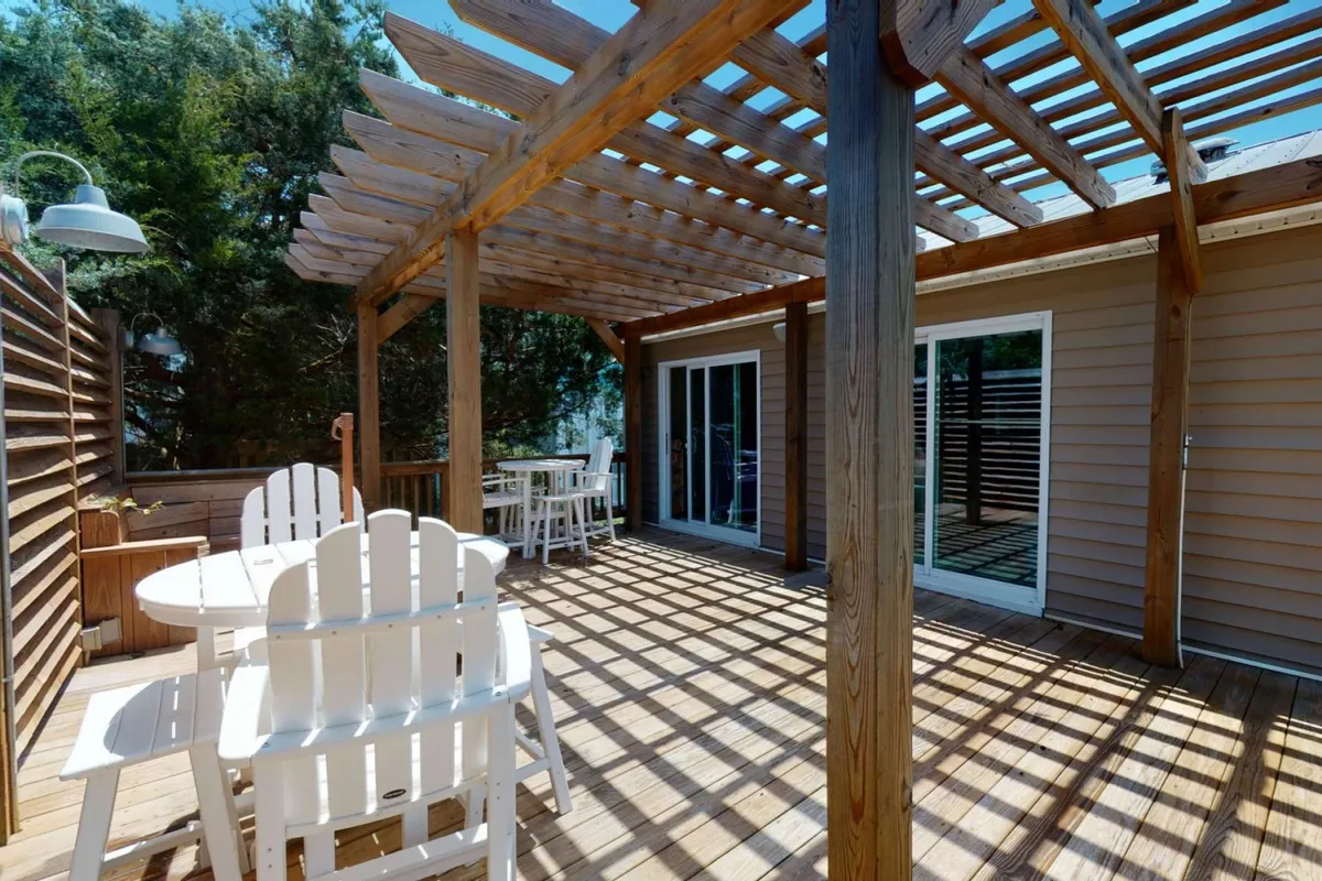 A stylish outdoor deck featuring a pergola, white lounge furniture, and glass sliding doors connecting to the house. The scene evokes the feel of a relaxed gathering space for enjoying cocktails, quiet conversations, or laid-back meals outdoors.