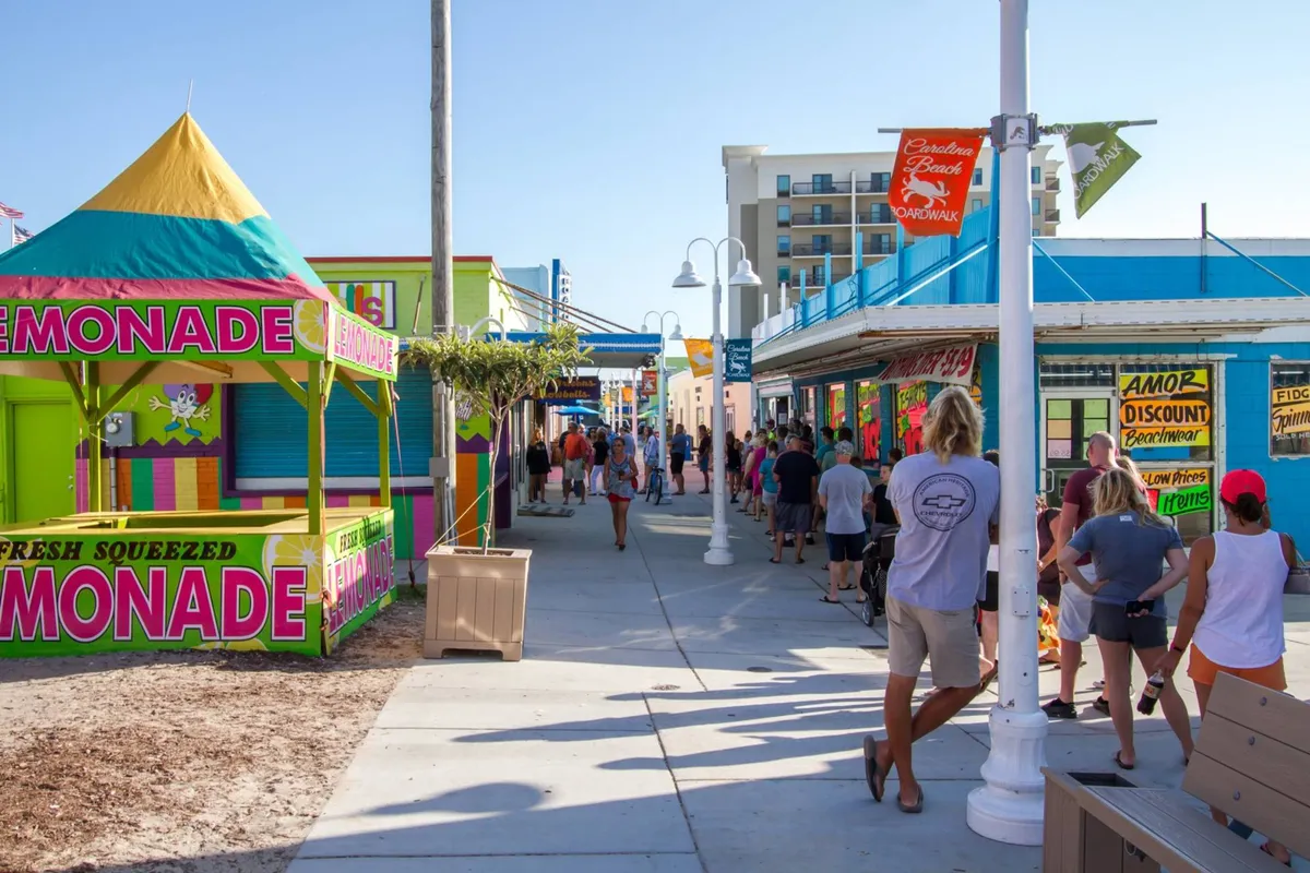 A lively scene at the Carolina Beach Boardwalk, featuring a colorful lemonade stand, cheerful crowds, and vibrant storefronts—capturing the energetic, festive ambiance of the area.