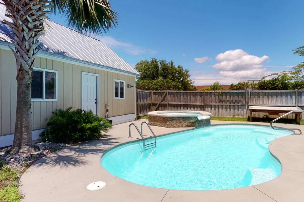 A tranquil backyard scene featuring a private swimming pool surrounded by lush greenery and a spa area under the shade of a tall palm tree. The pool area is bordered by a spacious deck with plenty of room for lounging, sunbathing, and enjoying gatherings under the evening sky.