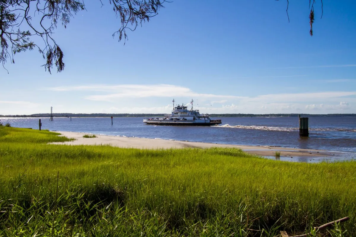 A serene view of a ferry gliding across tranquil water, framed by a grassy shoreline and distant trees. The scene invites thoughts of ferry rides, waterfront picnicking, and peaceful moments in nature—activities highlighted in the local exploration narrative.