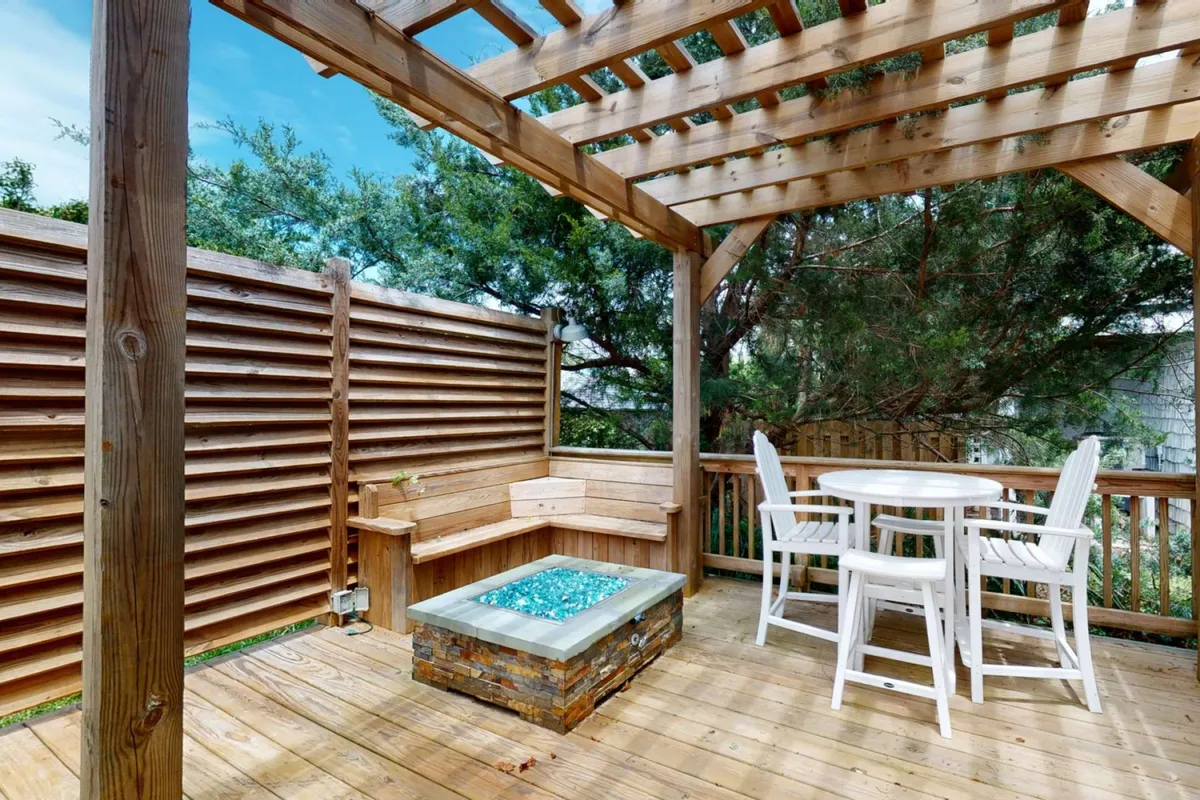 A cozy outdoor seating area arranged around a modern fire pit with blue glass stones, all beneath a pergola. The fire pit is surrounded by comfortable chairs, inviting guests to gather and enjoy the warmth under the evening sky.