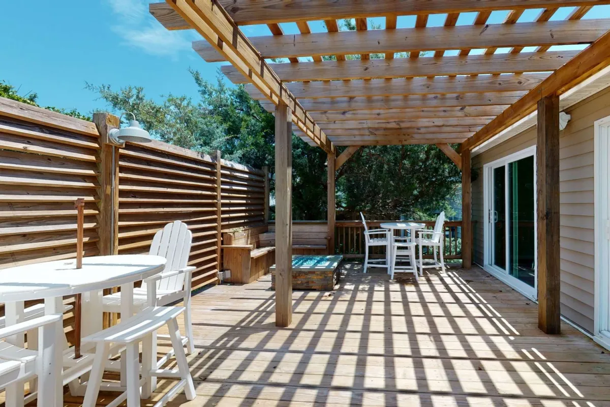 This image features a spacious wooden deck beneath a pergola, furnished with white chairs and a table. Lush greenery surrounds the area, creating an inviting spot for outdoor dining or gathering. The setup suggests a perfect environment for family breakfasts or game nights under the stars.