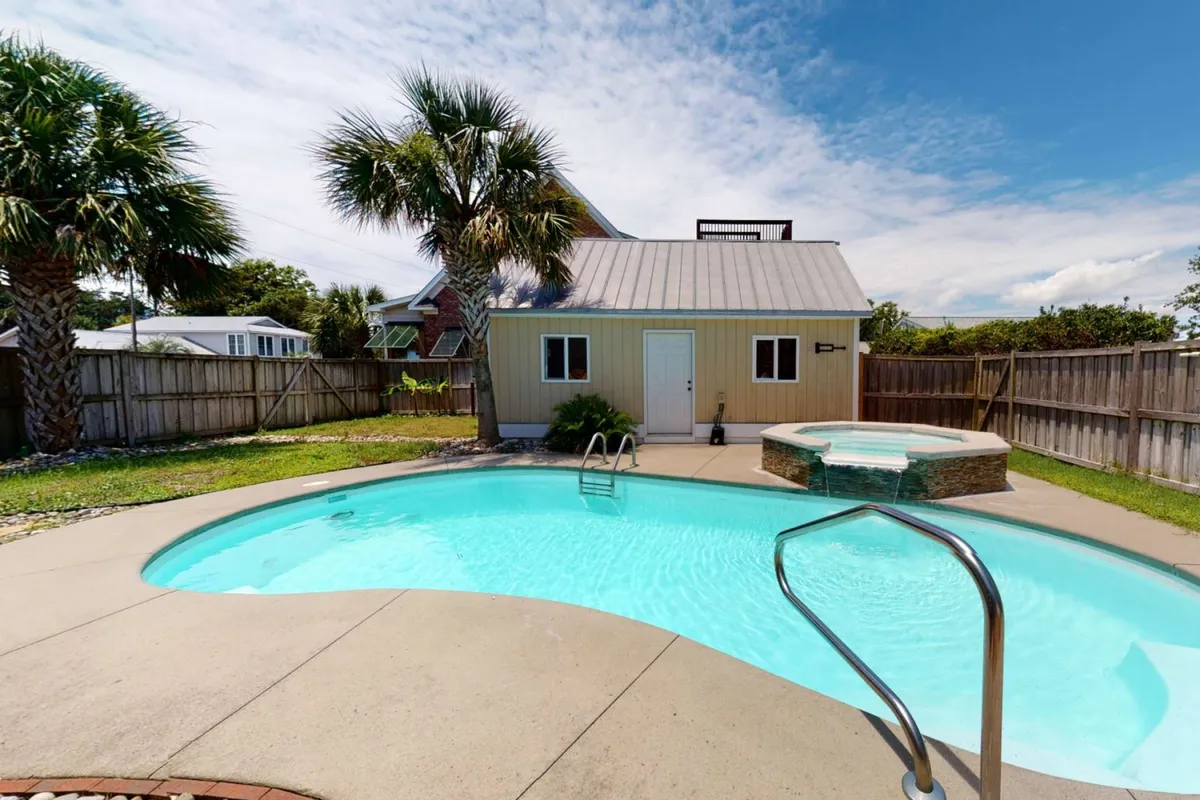The image showcases the exterior oasis of Tipsea Toes—a sparkling private swimming pool and inviting hot tub surrounded by lush palm trees, with a shed in the background. The scene is set for leisure, featuring ample space for sunbathing, barbecuing, and gathering with friends and family. The lighting suggests a tranquil, late afternoon or early evening, enhancing the sense of relaxation and retreat.