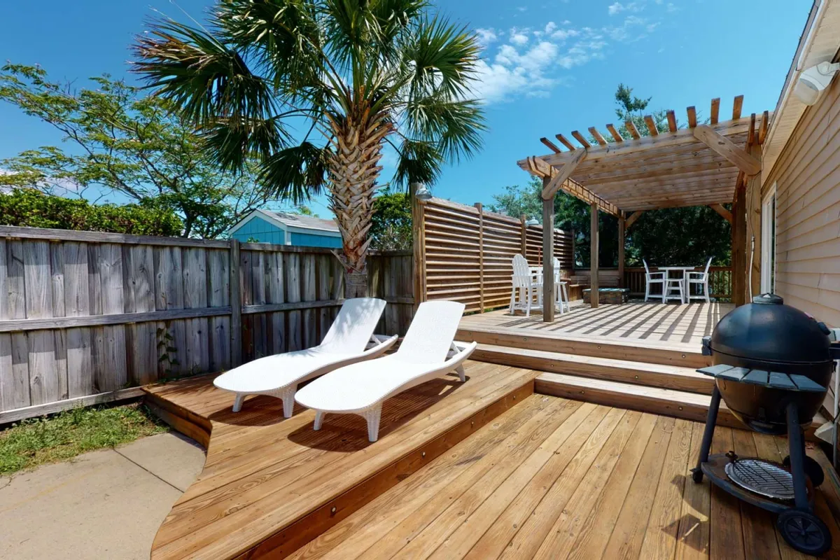 A wooden deck area furnished with lounge chairs and outdoor seating beneath a pergola, adjacent to a grill. The backdrop features palm trees, and the atmosphere is set for lazy afternoons or group barbecues as the daylight fades to sunset.