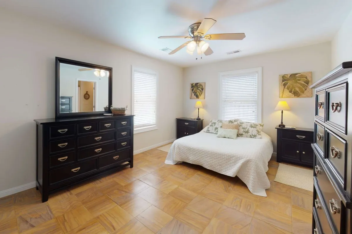 A peaceful bedroom featuring a queen bed with crisp white bedding, a dark wood dresser, a mirrored dresser, table lamps, and botanical wall art. Soft morning light filters into the space, creating a tranquil and restful atmosphere.
